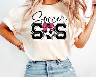 May include: Cream-colored t-shirt with the text "Soccer SIS" in black script and block letters. A soccer ball with a pink bow is centered between the letters "S". The shirt is paired with blue denim jeans.