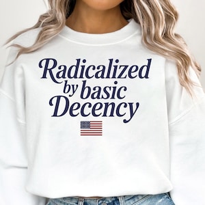 Puede incluir: Sudadera blanca con el texto "Radicalizado por la decencia básica" en letras azul marino. Una pequeña bandera estadounidense está impresa debajo del texto.