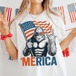 May include: White t-shirt featuring a graphic of a Bigfoot-like creature wearing sunglasses and holding an American flag. The word "'MERICA" is printed in orange and blue. The design is patriotic and suitable for Independence Day.