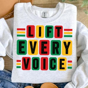 May include: A light gray sweatshirt with the words "LIFT EVERY VOICE" in black block letters. Each word is in a different colored square: red, yellow, and green. The sweatshirt has a crew neck and long sleeves.