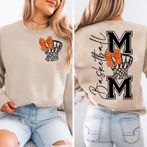 Puede incluir: Sudadera beige con un gr&aacute;fico de canasta de baloncesto y un lazo naranja. La parte trasera de la sudadera tiene las palabras "Basketball Mom" y dos grandes letras "M". Una sudadera de estilo casual.
