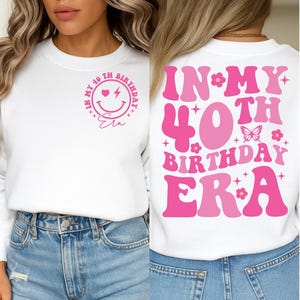 Può includere: Felpa bianca con testo e grafica rosa. La parte anteriore presenta una faccina sorridente, un cuore, un fulmine e le parole "In My 40th Birthday Era". Il retro ha le parole "In My 40th Birthday Era" con una farfalla e fiori.