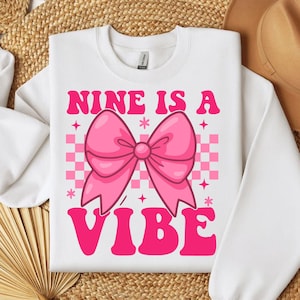 May include: White sweatshirt with the text "NINE IS A VIBE" in pink, featuring a large pink bow graphic. The background has a pink and white checkered pattern with star accents. A casual, fun design.