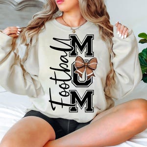 May include: Cream-colored sweatshirt with the text "Football Mom" in black script and block letters. A brown football bow is centered on the word "MOM". The sweatshirt is a casual, comfortable style.