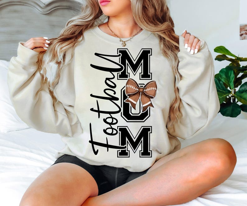 May include: Cream-colored sweatshirt with the text "Football Mom" in black script and block letters. A brown football bow is centered on the word "MOM". The sweatshirt is a casual, comfortable style.