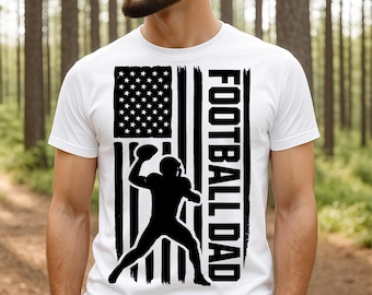 May include: White t-shirt featuring a black and white graphic of an American flag with a football player silhouette and the words "FOOTBALL DAD" in bold, vertical text. The shirt is a classic crew neck style.