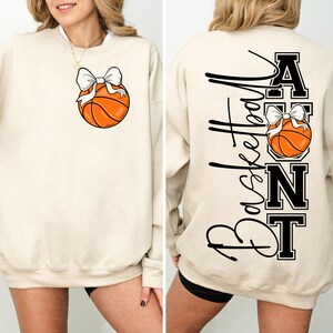 Op de afbeelding: Cr&egrave;mekleurige sweatshirt met een basketbal en een witte strik. De achterkant toont het woord "Basketball" in script, met "AUNT" in blokletters, en een andere basketbal met een strik.