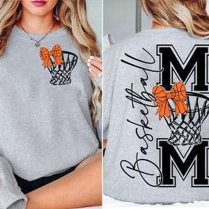 Puede incluir: Sudadera gris claro con una red de baloncesto y un lazo naranja. La parte trasera de la sudadera tiene la palabra "Basketball" en cursiva, con las letras "MOM" en negrita, en negro.