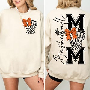 Puede incluir: Sudadera beige con una red de baloncesto y un lazo naranja. La parte trasera presenta las palabras "Basketball Mom" y las letras "MOM" en negro. Una prenda c&oacute;moda para los aficionados al baloncesto.