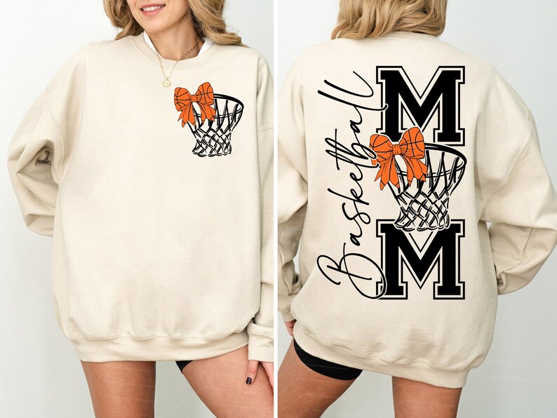 Puede incluir: Sudadera beige con una red de baloncesto y un lazo naranja. La parte trasera presenta las palabras "Basketball Mom" y las letras "MOM" en negro. Una prenda c&oacute;moda para los aficionados al baloncesto.