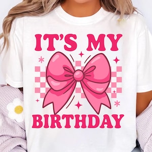 May include: White t-shirt with a pink bow graphic and the text "IT'S MY BIRTHDAY". The design includes a pink and white checkered background, stars, and floral accents. A fun and festive design.