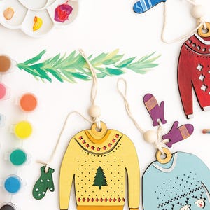 May include: Wooden Christmas sweater ornaments in red, yellow, and blue, with matching mitten accents. The ornaments are decorated with festive patterns and a Christmas tree. Paint and a paintbrush are also visible.