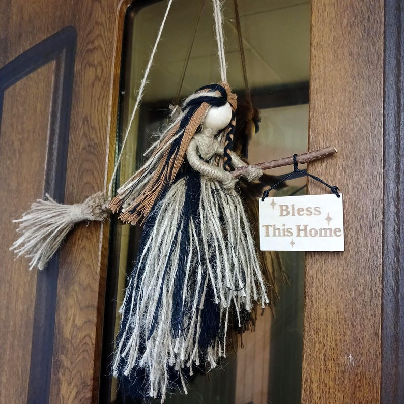 May include: A handmade witch doll with a broom and a sign that reads "Bless This Home." The doll is made of twine and yarn in black, brown, and cream colors. The sign is rectangular and has a light wood finish.