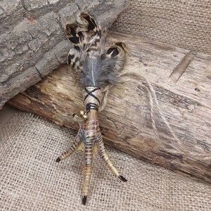 Op de afbeelding: Een decoratief item met een vogelpoot met bevestigde veren en kleine belletjes. De poot is omwikkeld met touw en zwart touw, en de veren zijn bruin, zwart en grijs. Een natuurlijke, rustieke uitstraling.