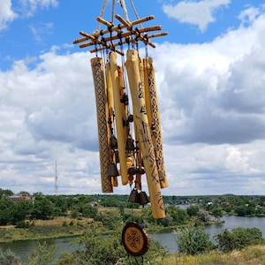 Pode incluir: Um carrilhão de vento de bambu com vários tubos ocos suspensos numa estrutura de madeira. Os tubos estão decorados com padrões geométricos e símbolos gravados em tons escuros. Pequenos sinos de metal e contas coloridas estão pendurados.