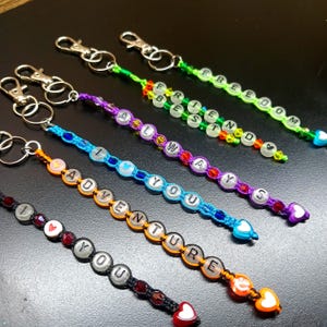 May include: Keychains with beaded designs and silver-tone clasps. The keychains feature phrases like "I LOVE YOU", "ADVENTURE", "ALWAYS", "FRIEND", and "FREEDOM". Each keychain has a heart-shaped charm at the end, with a variety of colors.