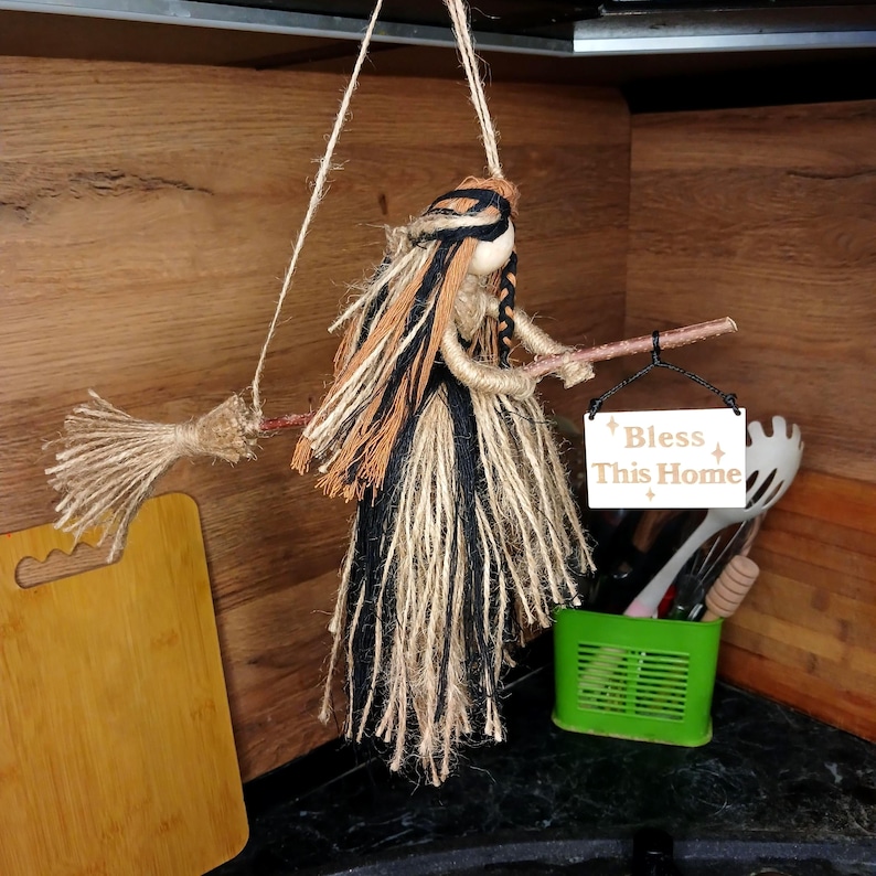 May include: A handmade witch doll with a broom, crafted from natural twine and yarn. The doll has black, brown, and beige hair and holds a sign that reads "Bless This Home". The witch is suspended by a string, suggesting it's a hanging decoration.