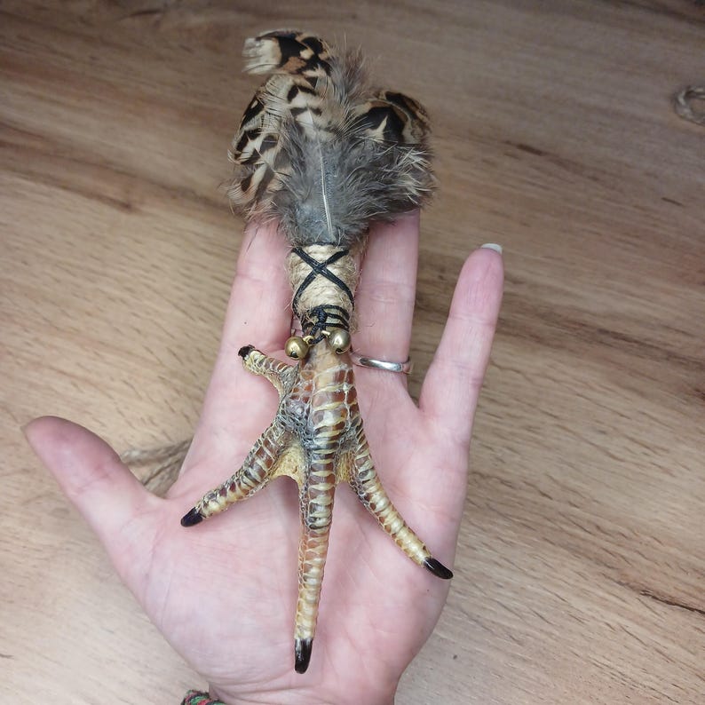 Op de afbeelding: Een unieke, handgemaakte bedel met een vogelpoot met veren en kleine belletjes. De poot is bruin en beige, met zwarte klauwen. De veren zijn bruin en grijs. De bedel is omwikkeld met touw.