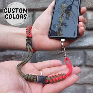 Op de afbeelding: Een rood en groen paracord telefoonkoord met een zwarte gesp en polsband. Het koord is bevestigd aan een zwarte smartphone. De tekst "CUSTOM COLORS" staat in een witte cirkel linksboven.