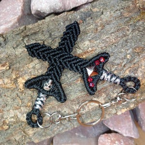 May include: Two black macrame dagger keychains with silver accents and skull and gem details. One dagger has red gems. The keychains are connected by a silver chain and key ring. The keychains are displayed on a piece of wood.