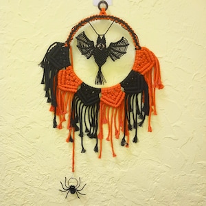 May include: A macrame Halloween decoration featuring a black bat pendant within a circular frame. The frame is adorned with alternating orange and black macrame squares and fringe. A small black spider hangs below.