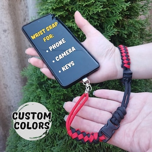 Op de afbeelding: Zwarte telefoon met een polsbandje van rood en zwart geweven koord. De band is ontworpen voor telefoons, camera's en sleutels. De tekst op de telefoon luidt "WRIST STRAP FOR: PHONE, CAMERA, KEYS" en "CUSTOM COLORS".