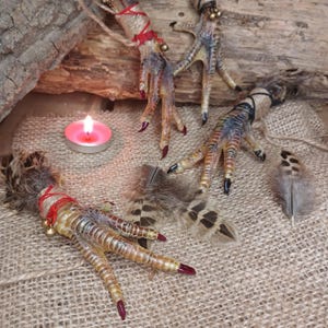 Op de afbeelding: Drie decoratieve vogelpoten met rood geschilderde klauwen, vastgebonden met rood touw en kleine gouden belletjes. De poten zijn gerangschikt met veren en een brandende roze kaars op een jute oppervlak.