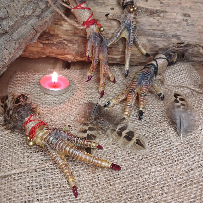 Op de afbeelding: Drie decoratieve vogelpoten met rood geschilderde klauwen, vastgebonden met rood touw en kleine gouden belletjes. De poten zijn gerangschikt met veren en een brandende roze kaars op een jute oppervlak.