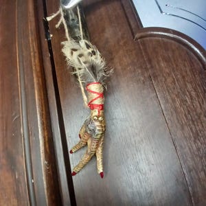 Op de afbeelding: Een decoratieve hanger met een vogelpoot met rood geschilderde klauwen, omwikkeld met touw en rood lint. De hanger bevat veren en wordt opgehangen aan een natuurlijke vezelkoord, hangend aan een deurklink.