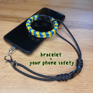 May include: A black smartphone with a blue and yellow braided bracelet attached, connected to a black lanyard with a clasp. The text "bracelet + your phone safety" is visible.