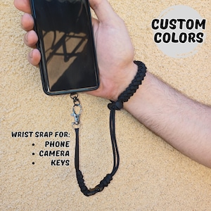 Op de afbeelding: Zwarte polsband voor telefoon, camera of sleutels. De band is bevestigd aan een telefoon en heeft een gevlochten polsband en een zilverkleurige sluiting. Tekst op de afbeelding luidt "Custom Colors" en "Wrist strap for: Phone, Camera, Keys."