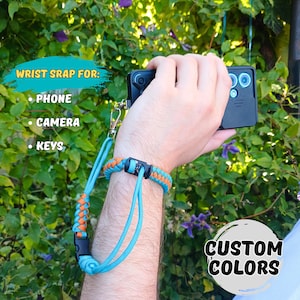Op de afbeelding: Een turquoise en oranje polsband met een zwarte sluiting is bevestigd aan een telefoon. De band is ontworpen voor telefoons, camera's en sleutels. De tekst "Custom Colors" is zichtbaar in een witte cirkel.