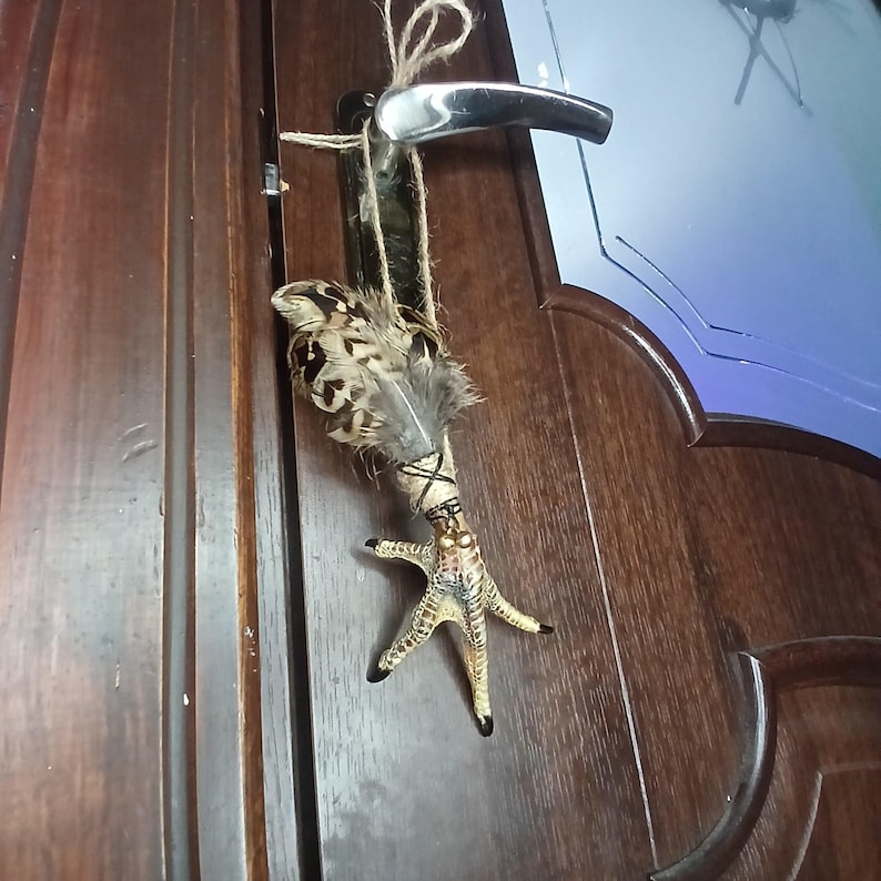 Op de afbeelding: Een decoratieve hanger met een vogelpoot met zwarte klauwen, versierd met veren en touw. De hanger hangt aan een deurklink met een natuurlijke touwlus. De veren hebben een patroon van bruin, zwart en cr&egrave;me.