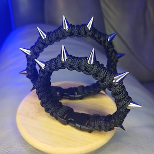 Könnte beinhalten: Zwei schwarze Seilarmbänder mit silberfarbenen Metallspitzen. Die Armbänder sind mit schwarzen Kunststoffschnallen befestigt und liegen auf einer hellbraunen Holzoberfläche. Punk-Rock-Stil.