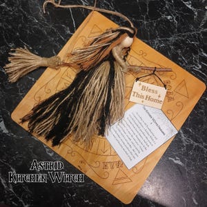 A handmade Norse kitchen witch doll in a black and tan dress, holding a miniature witch broom and a small personalized wooden sign, designed as a pagan good luck charm.