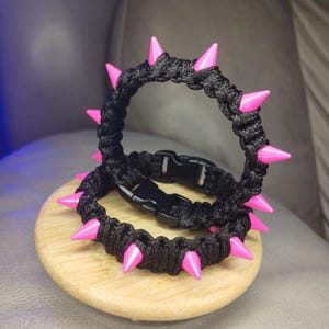 May include: Two black woven bracelets with bright pink spikes. Each bracelet has a black plastic buckle closure. The bracelets are displayed on a small, round wooden surface.