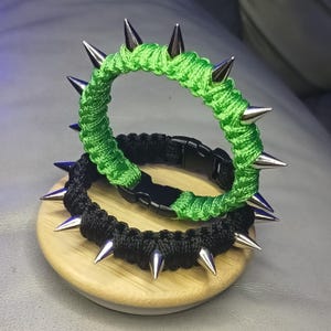 Może przedstawiać: Dwie bransoletki z kolcami. Jedna jest czarna ze srebrnymi kolcami, druga jasnozielona ze srebrnymi kolcami. Bransoletki wykonane są z tkanego materiału i leżą na małej drewnianej powierzchni.