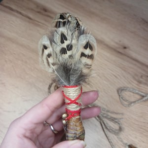 Op de afbeelding: Een decoratieve toverstaf met bruine en beige gespikkelde veren. De veren zijn bevestigd aan een basis gewikkeld met touw en rood koord. Kleine gouden belletjes hangen aan de basis en een stuk touw loopt uit van de toverstaf.