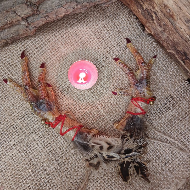 Op de afbeelding: Twee kippenpoten met rode accenten en kleine belletjes, gebonden met rood touw. Een kleine roze kaars staat tussen de poten. De poten liggen op een jute oppervlak.