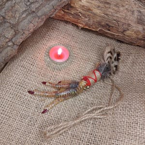 Op de afbeelding: Een gedroogde vogelpoot, versierd met veren, rood touw en een klein belletje, ligt op een jute oppervlak. De klauwen zijn rood geverfd. Een brandende roze kaars staat op de achtergrond.