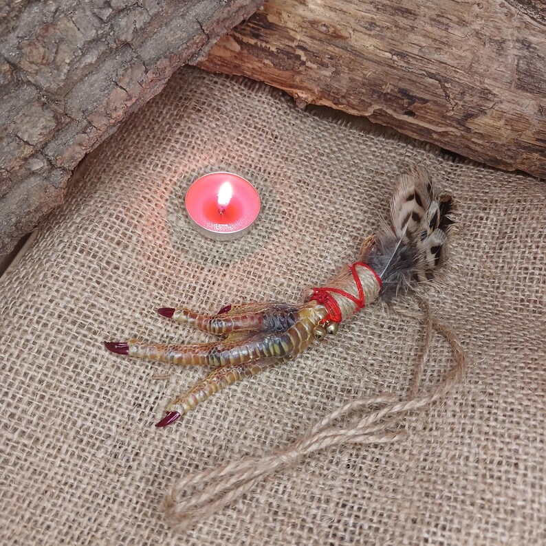 Op de afbeelding: Een gedroogde vogelpoot, versierd met veren, rood touw en een klein belletje, ligt op een jute oppervlak. De klauwen zijn rood geverfd. Een brandende roze kaars staat op de achtergrond.