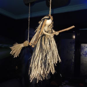May include: A handmade witch doll crafted from natural twine, featuring a wooden head and a broom. The witch's hair and dress are made of black and tan twine. The doll is suspended by twine, creating a whimsical, rustic aesthetic.