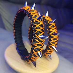 May include: Two black and orange paracord bracelets with silver spikes. The bracelets are woven with a criss-cross pattern.