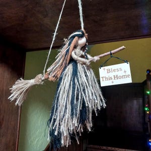 May include: A handmade witch doll with a broom, hanging from a string. The doll has a yarn dress in black, white, and blue, with brown and black hair. It holds a small sign that reads "Bless This Home".