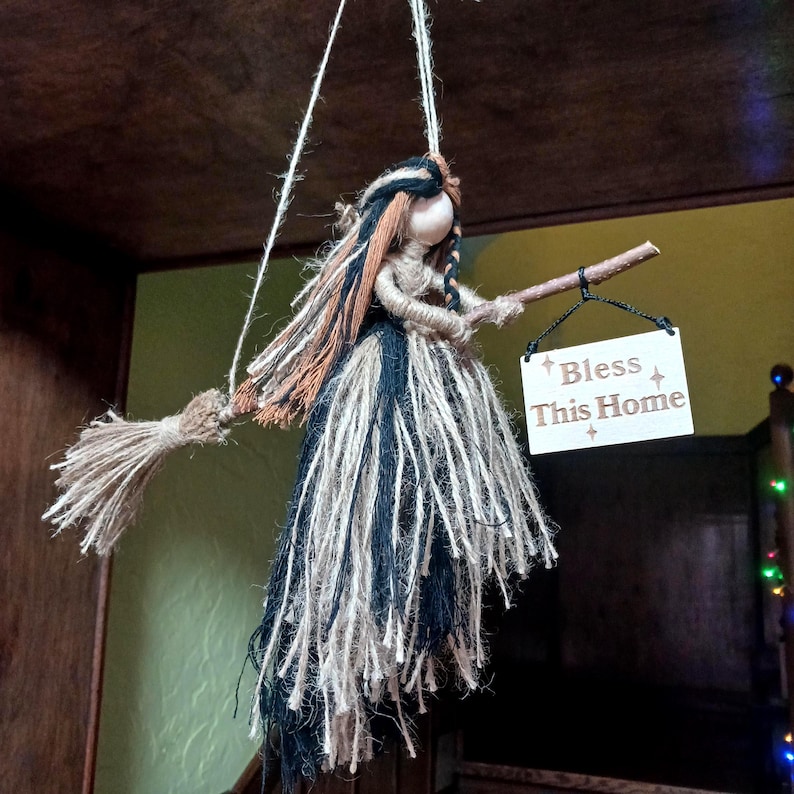 May include: A handmade witch doll with a broom, hanging from a string. The doll has a yarn dress in black, white, and blue, with brown and black hair. It holds a small sign that reads "Bless This Home".