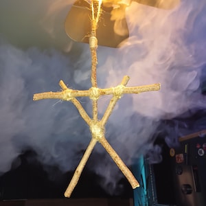 May include: A hanging ornament constructed from five brown wooden twigs arranged in a stick figure shape. The twigs are securely bound together at the joints with light tan twine, and a loop of the same twine extends from the top for hanging.