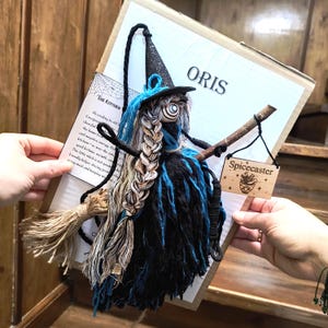 Peut inclure: Une poupée sorcière artisanale en fil noir et bleu, dotée d'un chapeau pointu noir pailleté, d'une coiffure tressée et d'un manche à balai en bois. Une petite pancarte en bois attachée au balai porte l'inscription Spicecaster sur une carte.