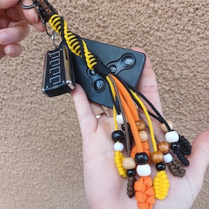 May include: A handmade keychain with a black, yellow, and orange braided cord design. The keychain features wooden and white beads, and is attached to a car key fob. The design includes a metal keyring.