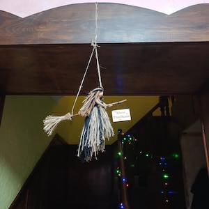 May include: A hanging witch decoration made of natural fibers, with a broom and a sign that reads "Bless This Home." The witch has orange, black, and tan hair and a long, flowing skirt. The decoration is suspended from a wooden beam.