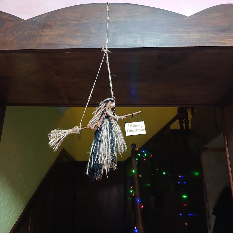 May include: A hanging witch decoration made of natural fibers, with a broom and a sign that reads "Bless This Home." The witch has orange, black, and tan hair and a long, flowing skirt. The decoration is suspended from a wooden beam.